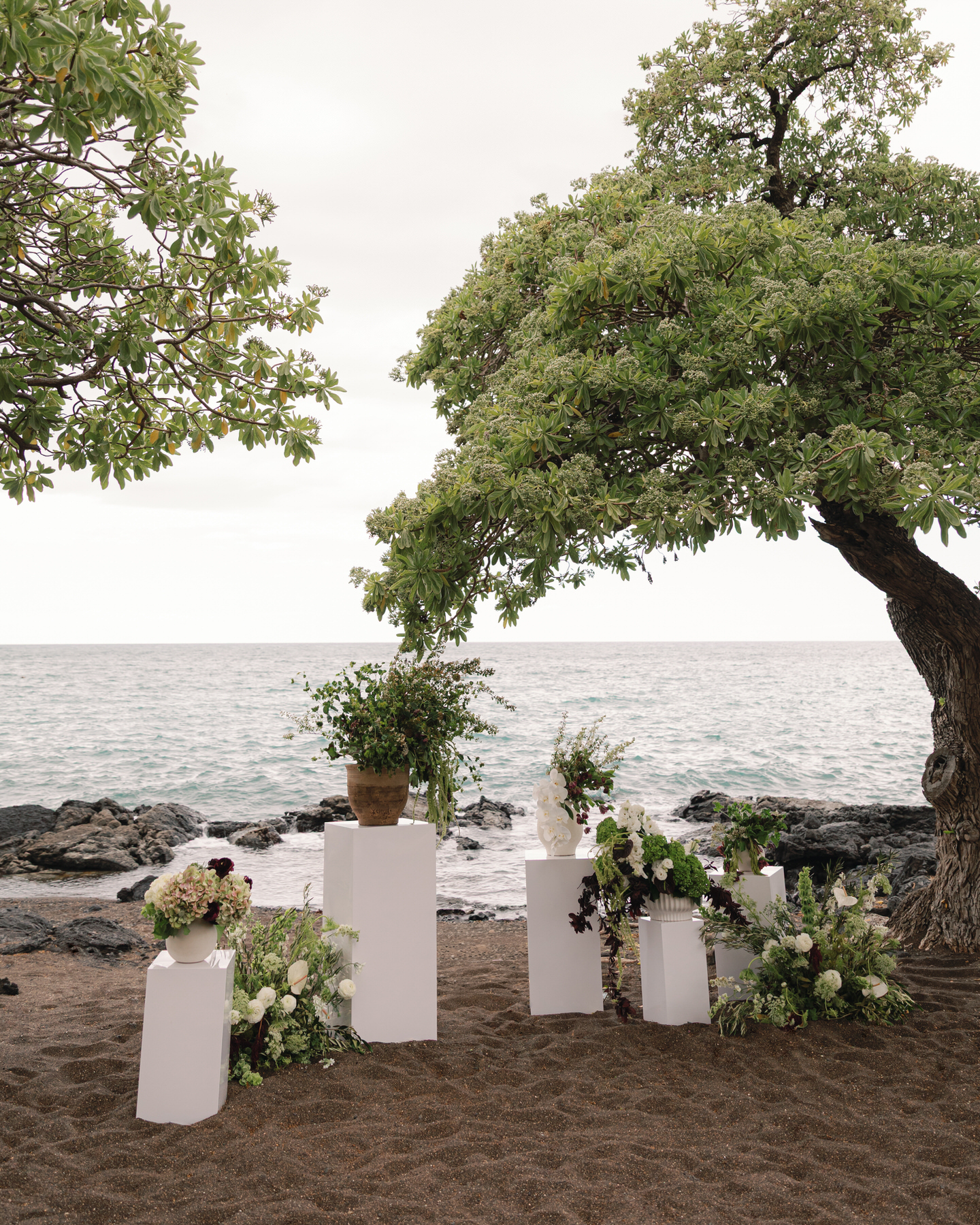A scenic view of a wedding setup by the beach in Oahu, with white pedestal tables holding lush flower arrangements. The natural beauty of the landscape, with a tree arching above the scene and the ocean in the distance, showcases the importance of planning early and hiring your Oahu wedding