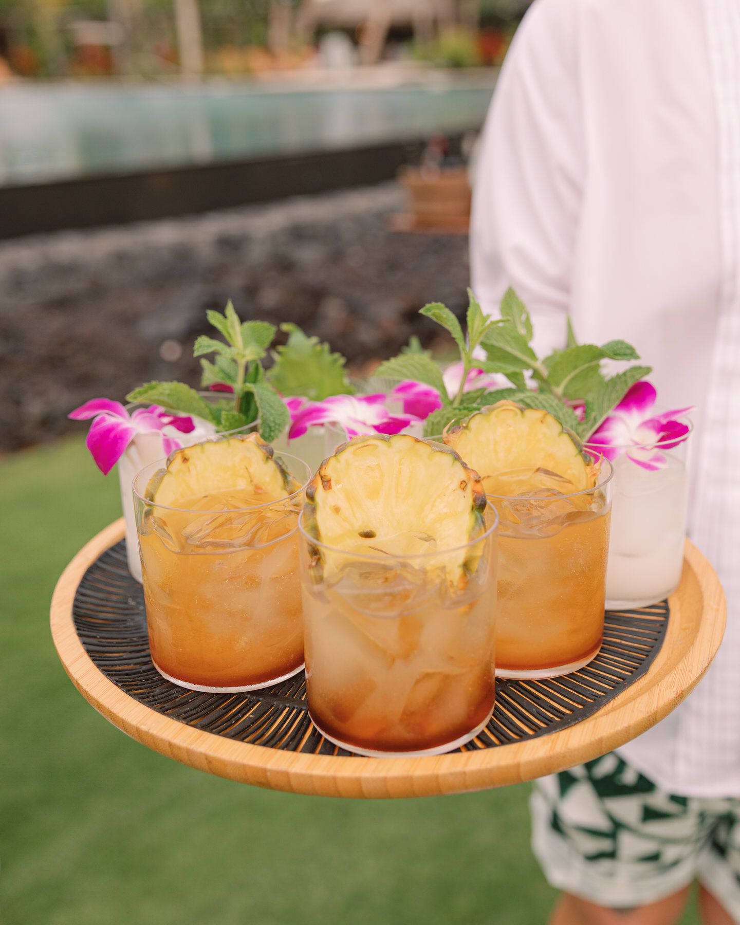 A tray of tropical cocktails, beautifully garnished with fresh pineapple and purple orchids, is presented at an outdoor wedding reception in Oahu. This refreshing drink selection highlights why it’s crucial to hire your Oahu wedding vendors ahead of time to ensure your guests experience personalized and unique offerings (and my personal recommendations).