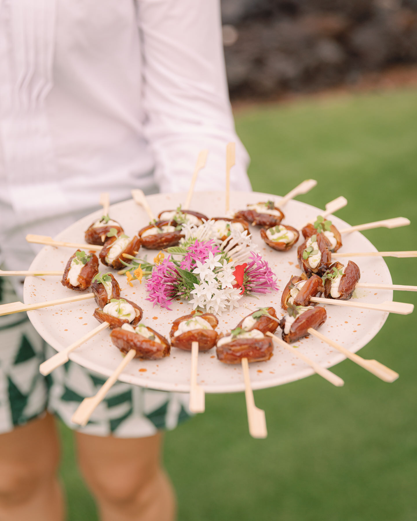 A server presents a beautifully arranged platter of appetizers, featuring date skewers garnished with herbs and edible flowers. This delightful dish exemplifies the importance of hiring your Oahu wedding vendors in advance to ensure that the catering at your wedding offers unique and memorable experiences (and my personal recommendations).