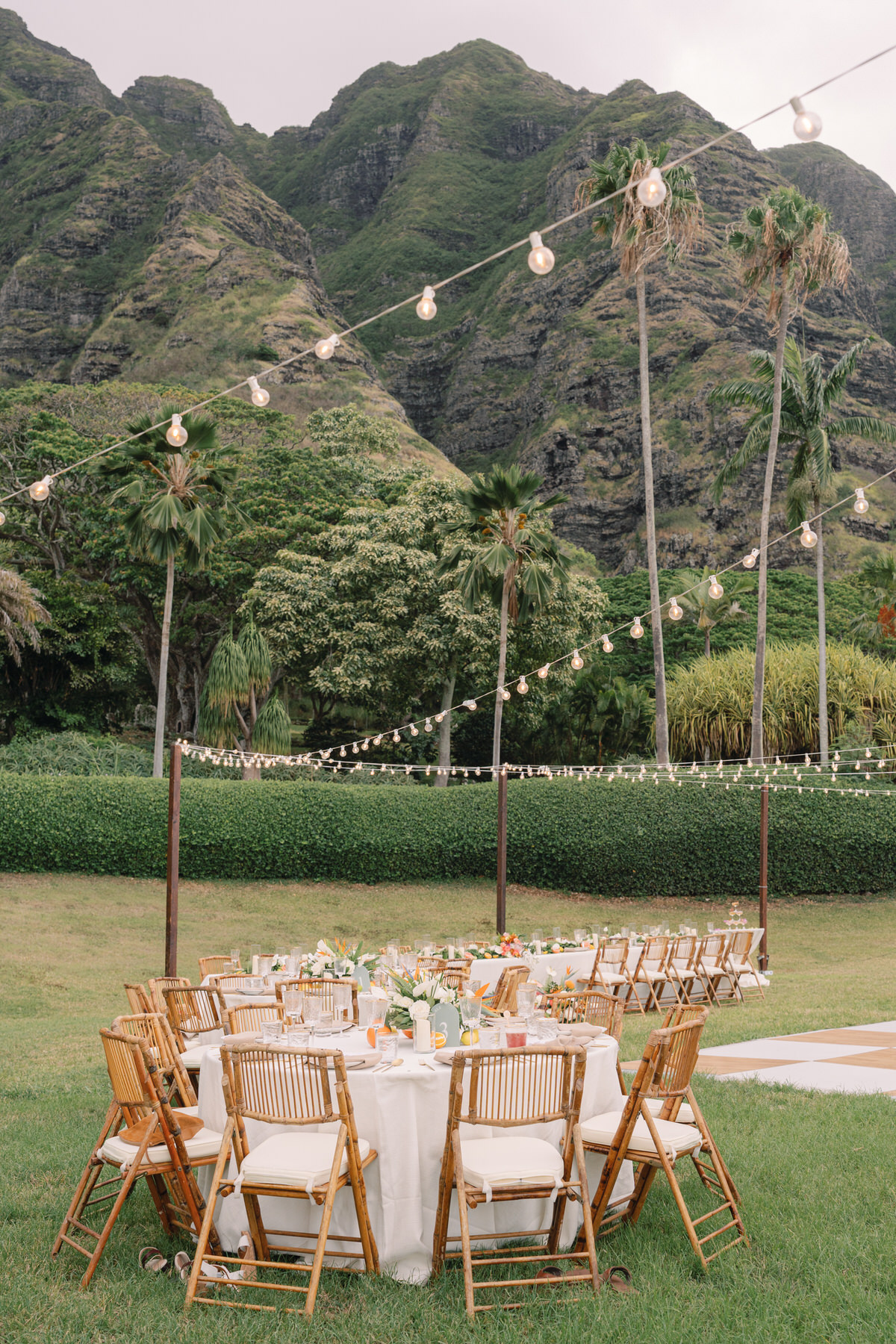 An elegant outdoor wedding reception setup with round tables dressed in white linens, surrounded by golden chairs. The string lights above add a warm glow to the scene, framed by the lush green mountains in the background. This idyllic setting emphasizes the importance of securing your Oahu wedding vendors early to ensure a flawless reception (and my personal recommendations).