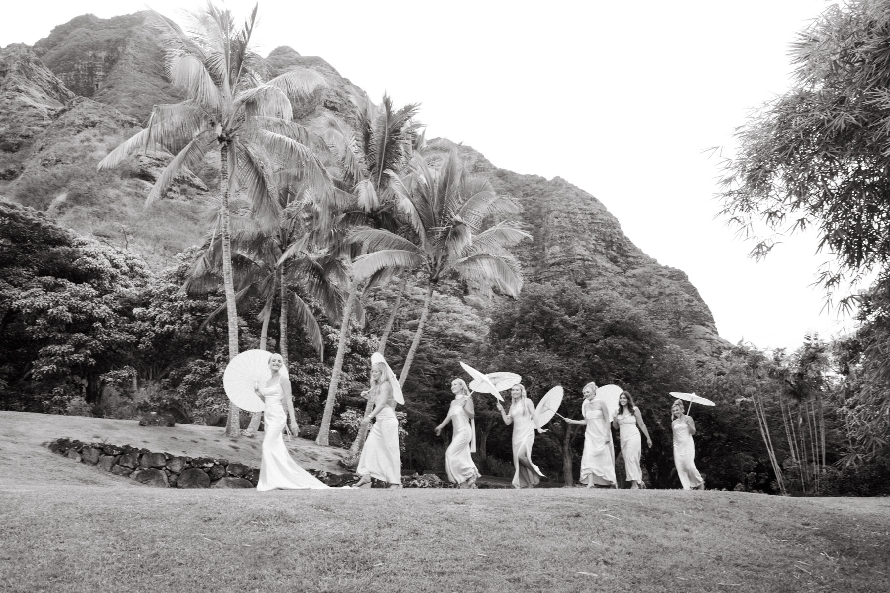 Bridesmaids walking with parasols in black and white at Kualoa Ranch, Paliku Gardens, Oahu, framed by stunning mountain scenery.