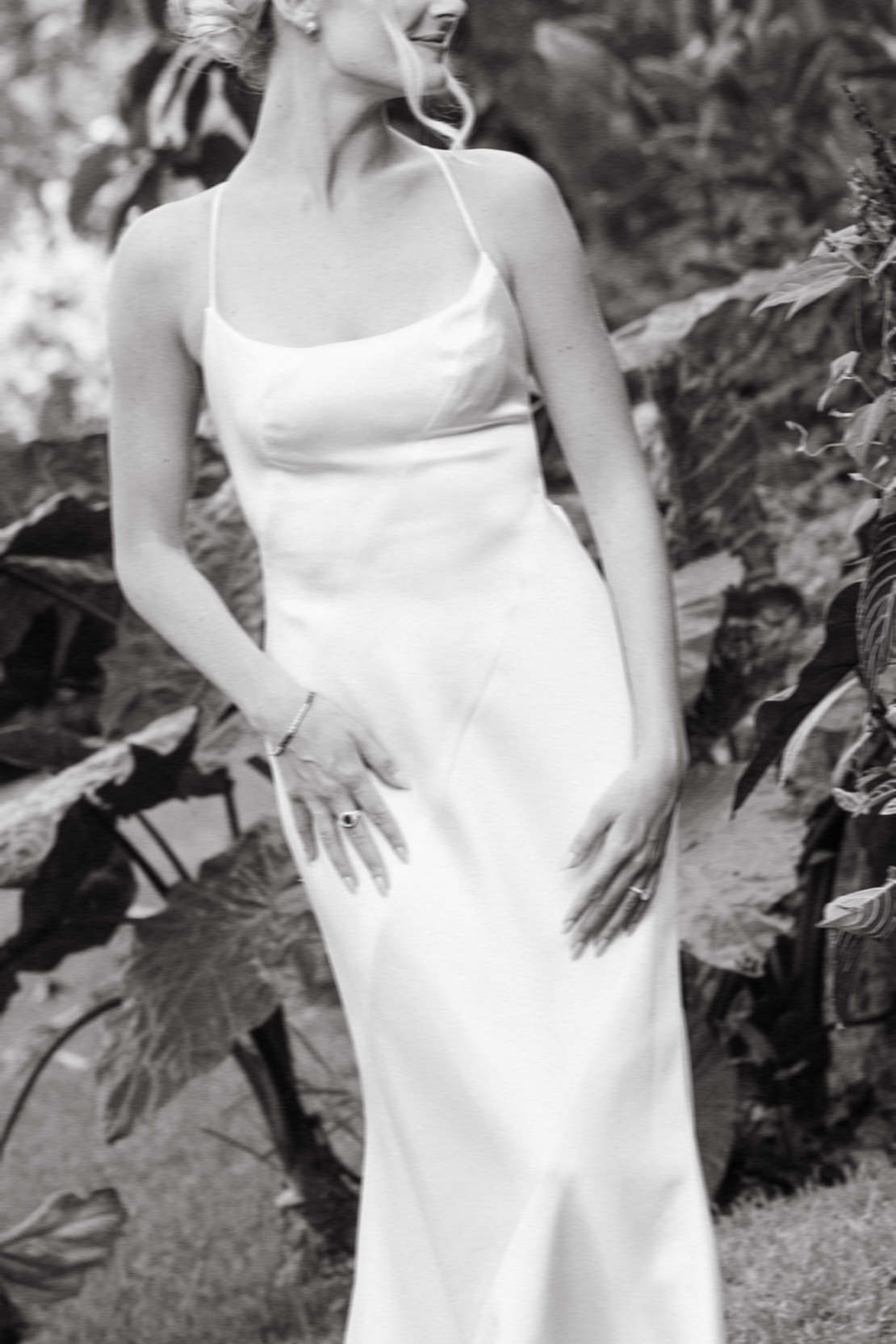 Bride in a simple white gown, with her hands resting on her dress, captured in black and white at Kualoa Ranch, Paliku Gardens.