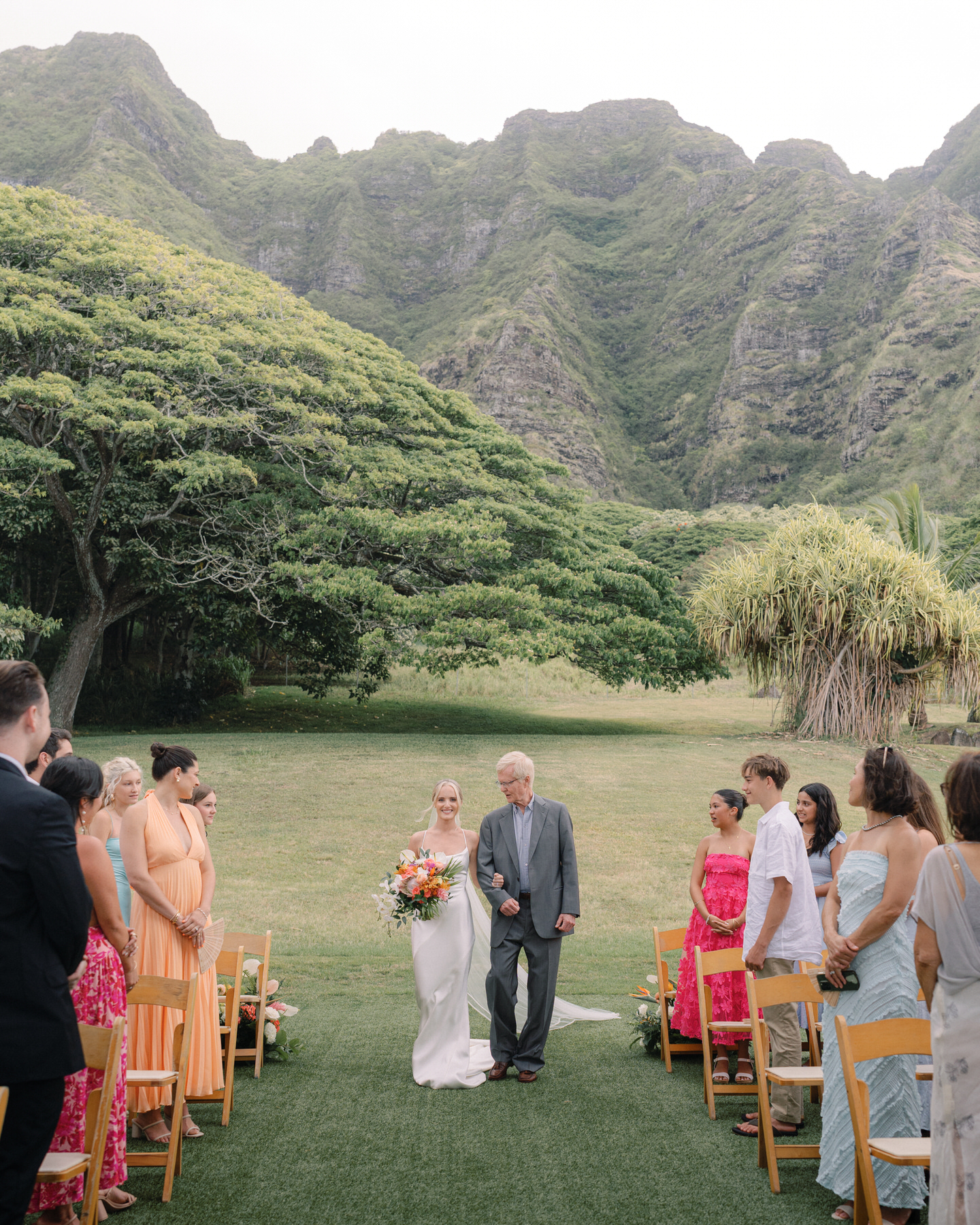 Bride walking down the aisle with her father at Kualoa Ranch, Paliku Gardens, Oahu, with dramatic mountain backdrop.