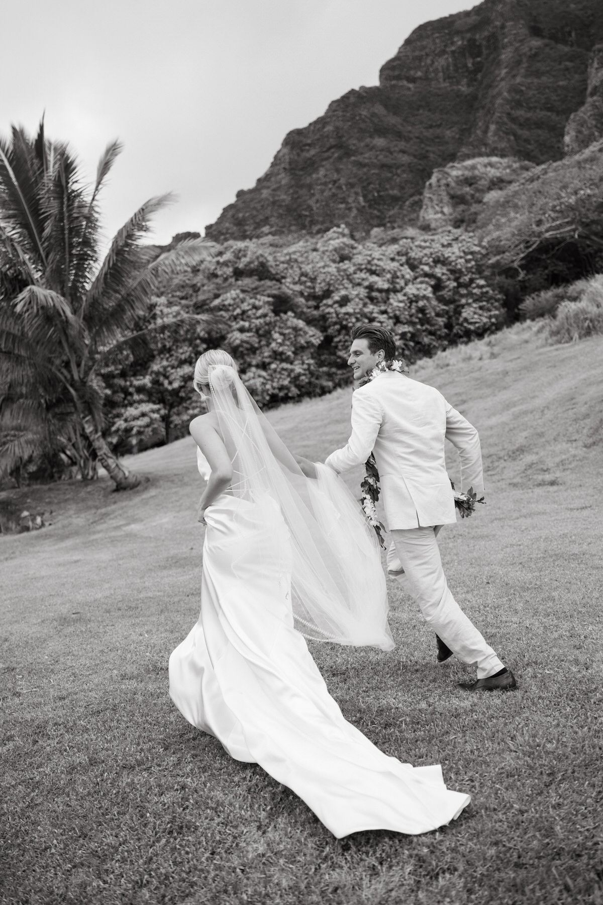 Bride and groom walking hand-in-hand under the palms, the groom leading with her veil flowing behind, captured at Kualoa Ranch, Paliku Gardens.