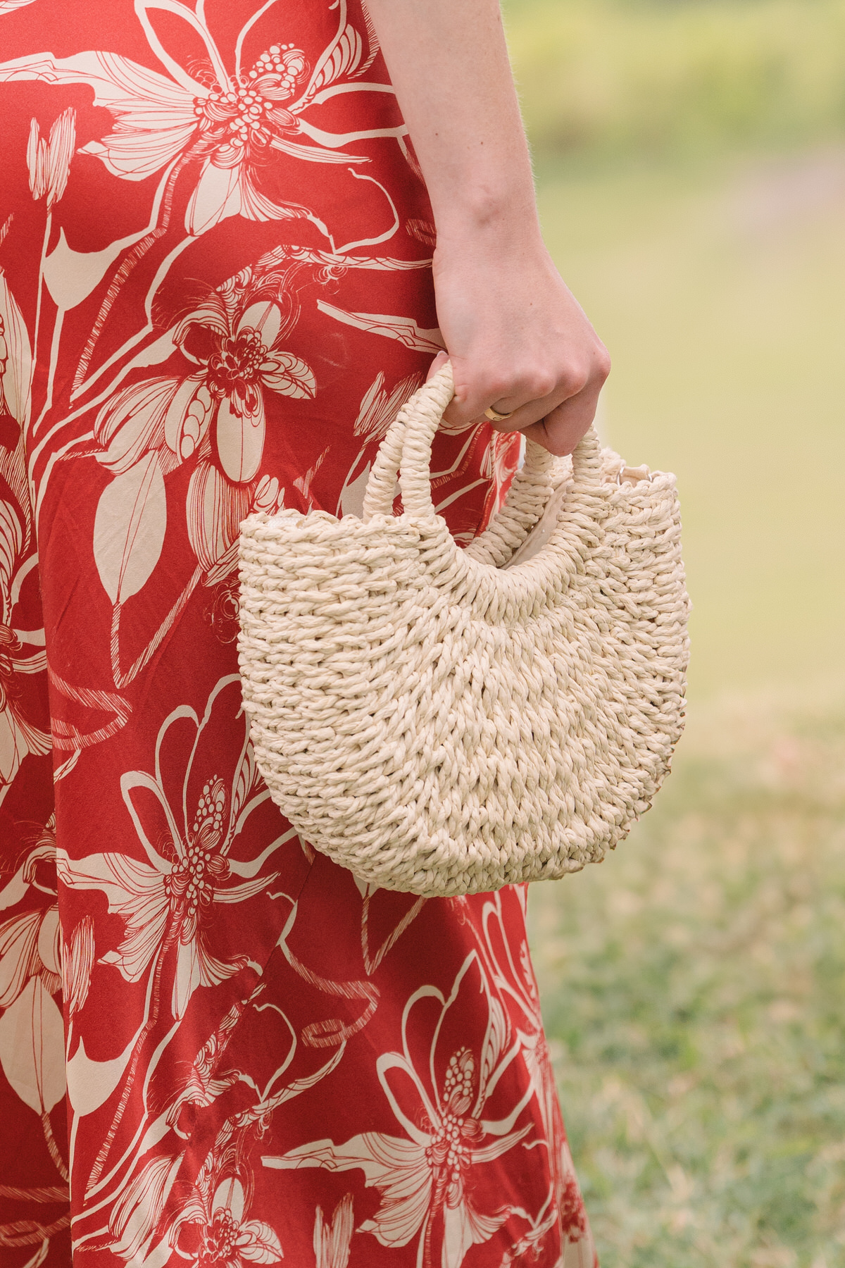 Close-up of a guest holding a woven handbag, with a floral red dress, at Kualoa Ranch, Paliku Gardens, during a wedding.