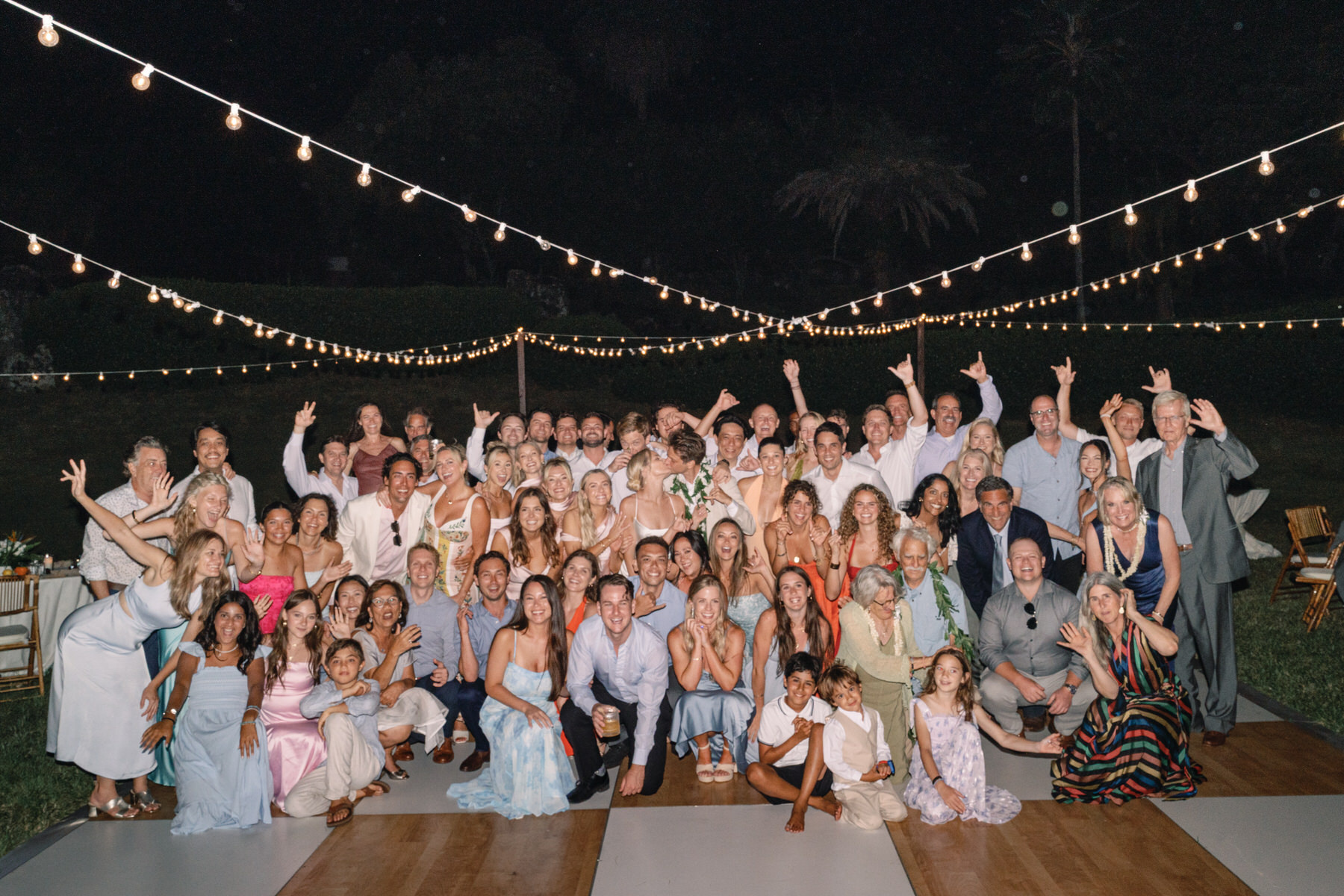 Large group photo of wedding guests celebrating on the dance floor at Kualoa Ranch, Paliku Gardens, with string lights above.