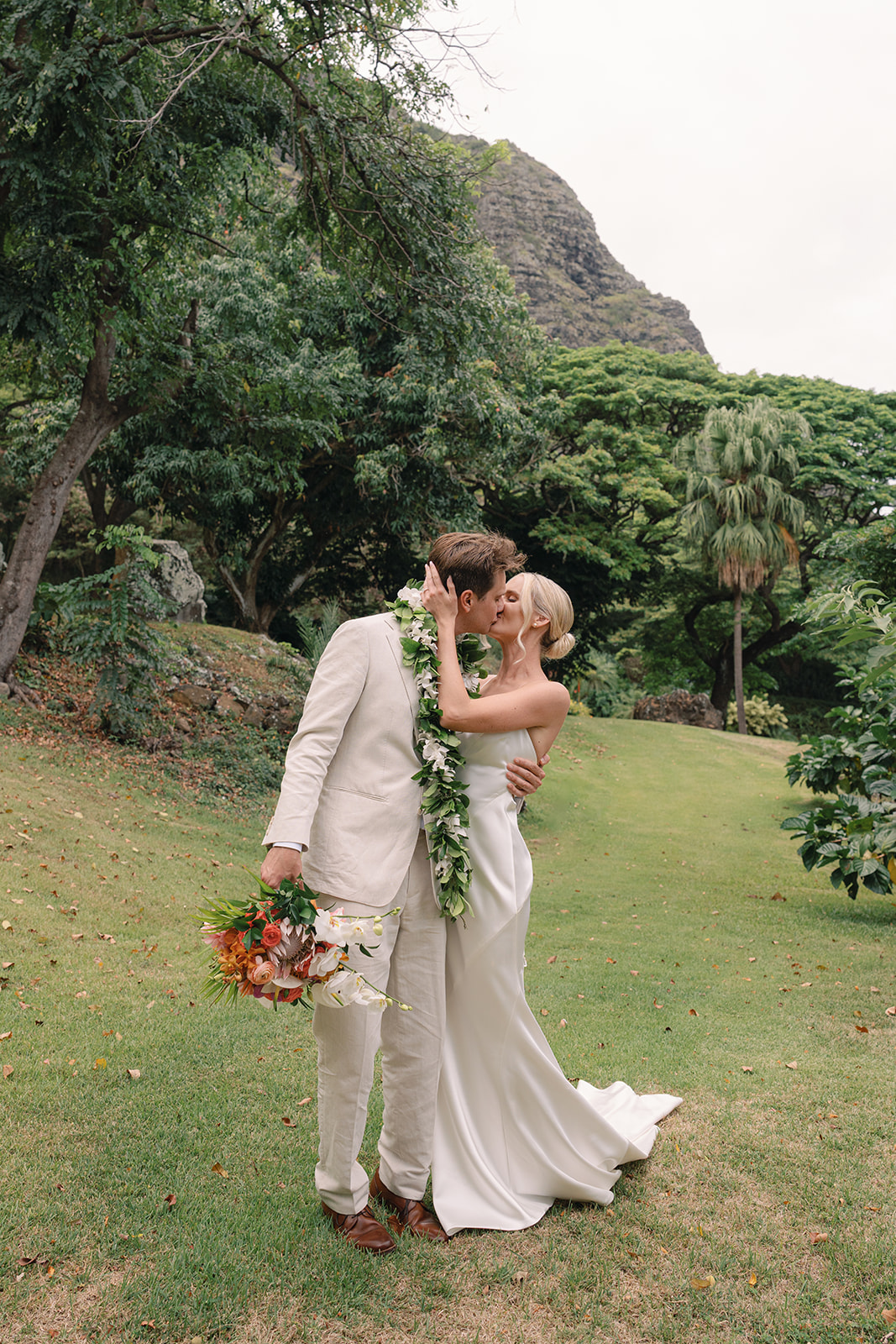 Bride and groom sharing a romantic kiss at Kualoa Ranch, Paliku Gardens, with lush tropical scenery in the background.