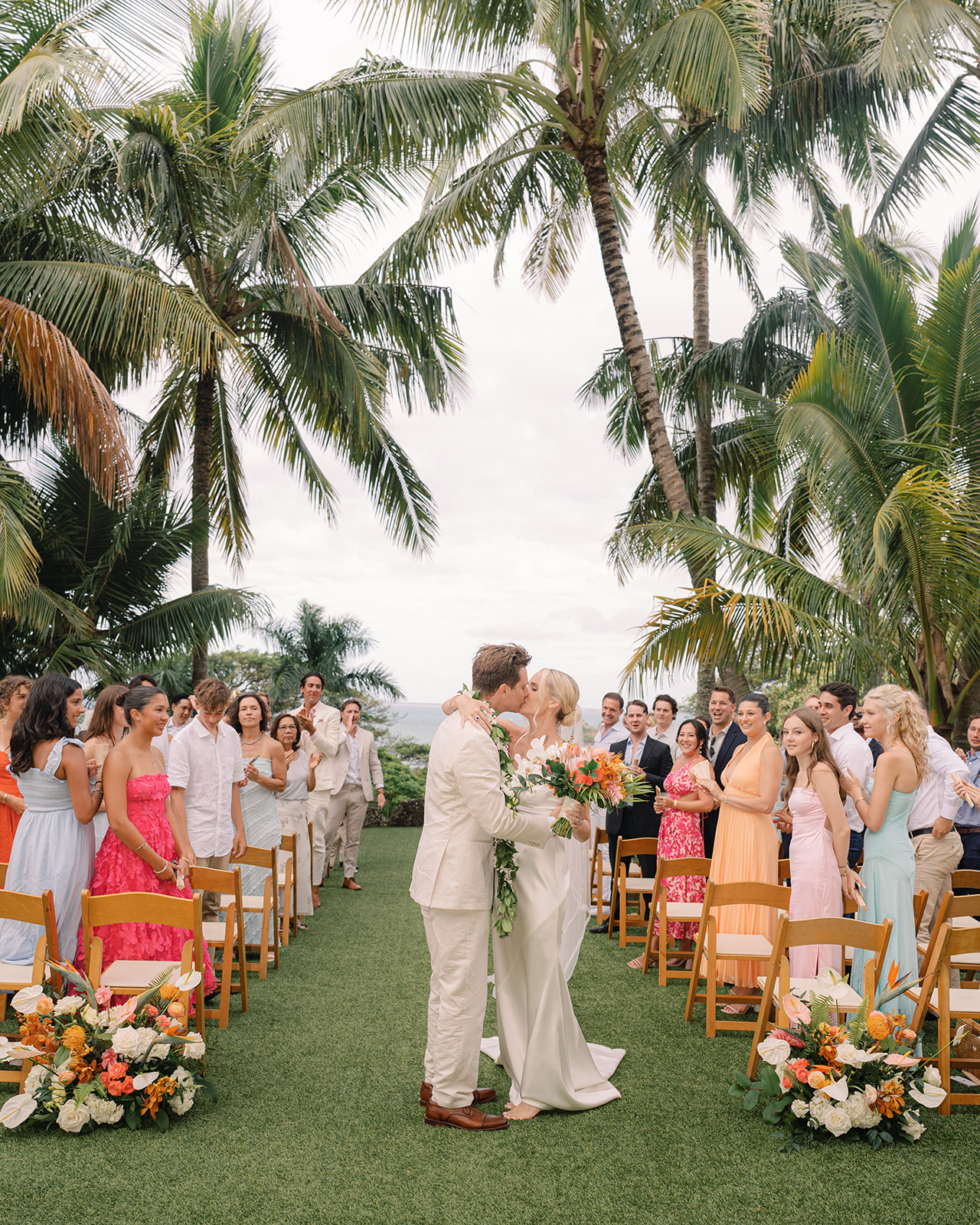 Couple sharing a kiss during their wedding ceremony at Kualoa Ranch, Paliku Gardens, Oahu, with tropical trees and guests in attendance.