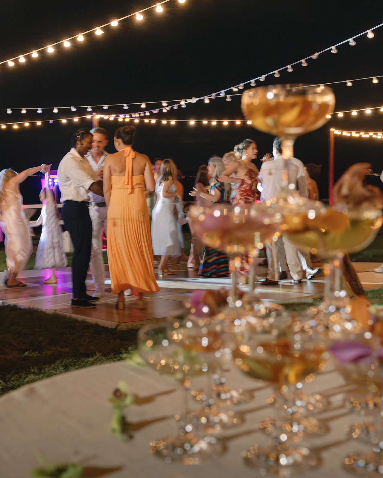 Wedding reception with champagne glasses in the foreground, guests dancing under string lights at Kualoa Ranch, Paliku Gardens