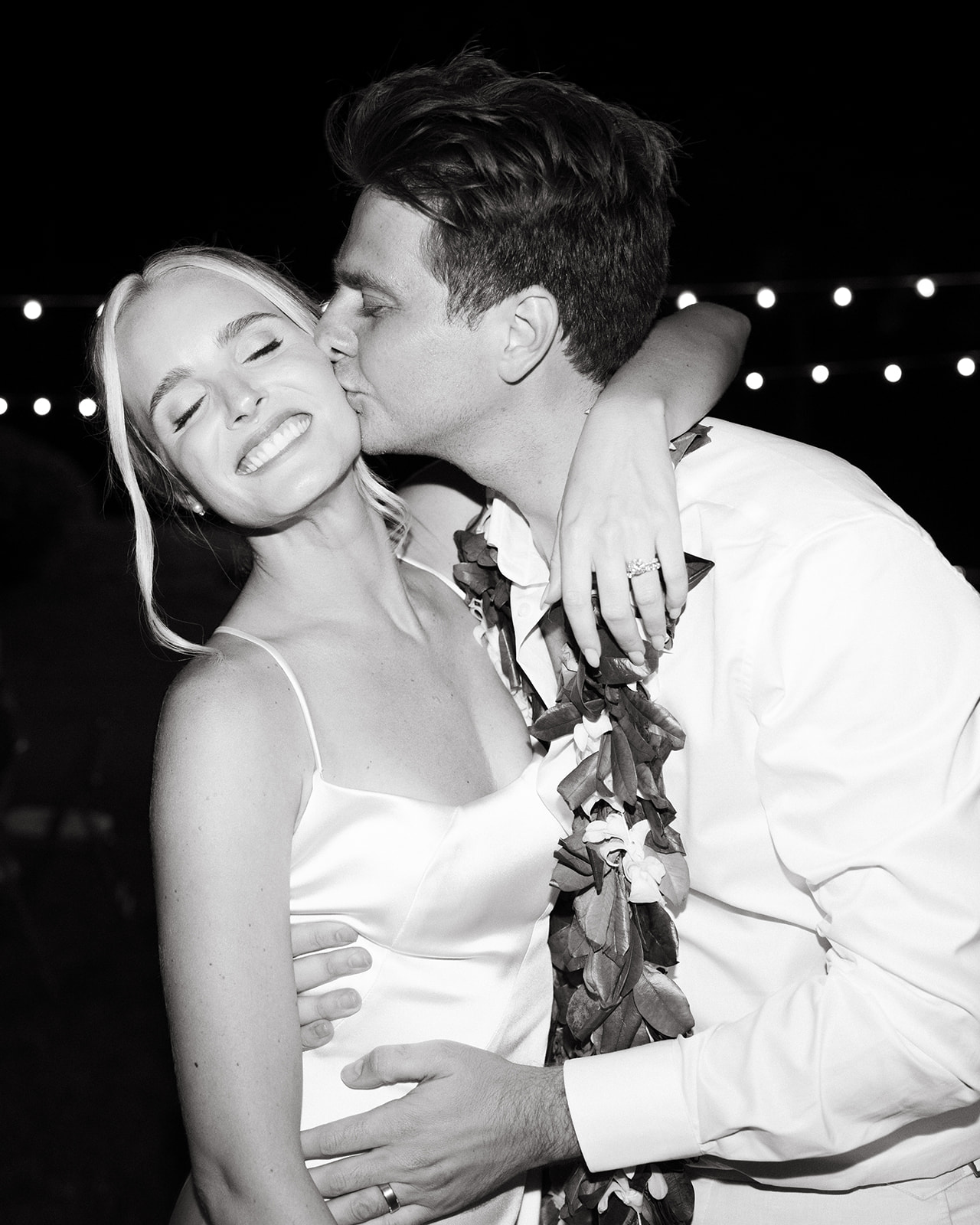 Bride and groom sharing a kiss at Kualoa Ranch, Paliku Gardens, captured in black and white with string lights in the background.