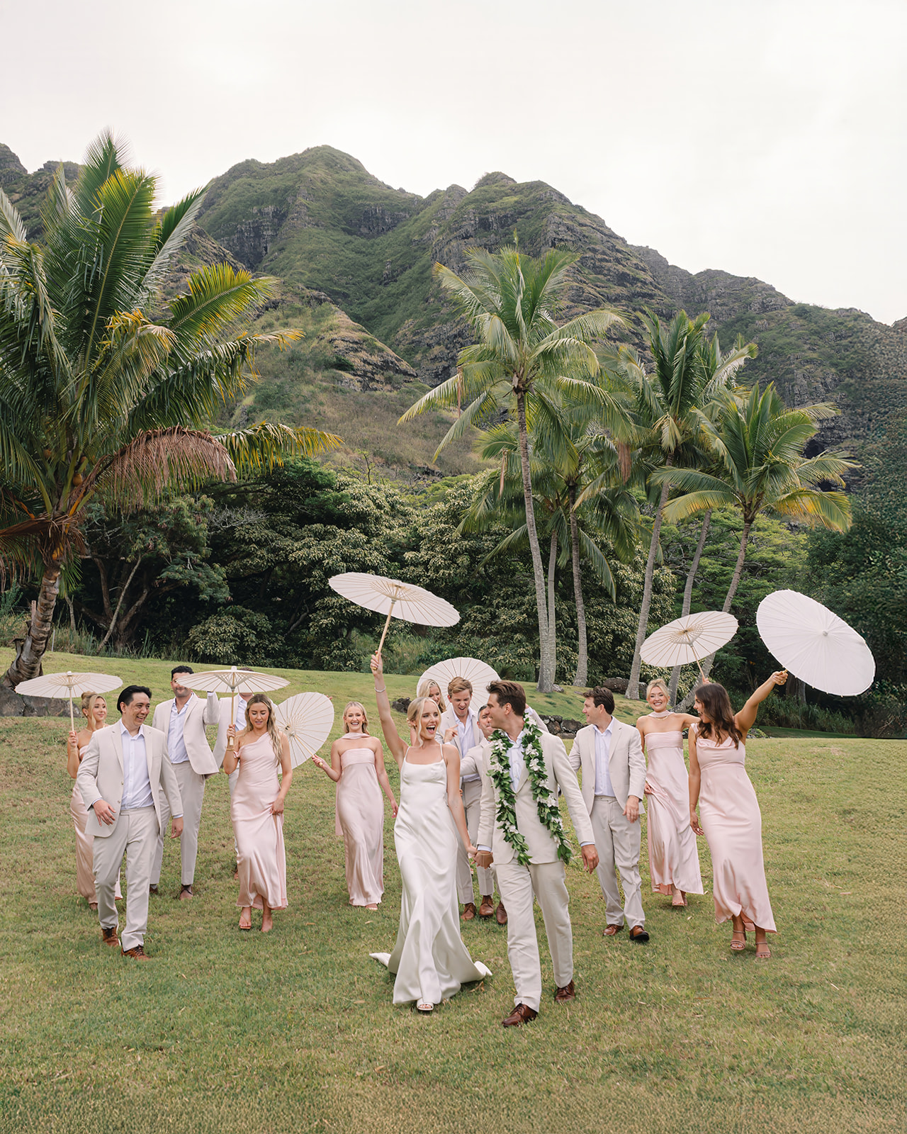 Bridal party celebrating with parasols at Kualoa Ranch, Paliku Gardens in Oahu, surrounded by lush tropical landscape.