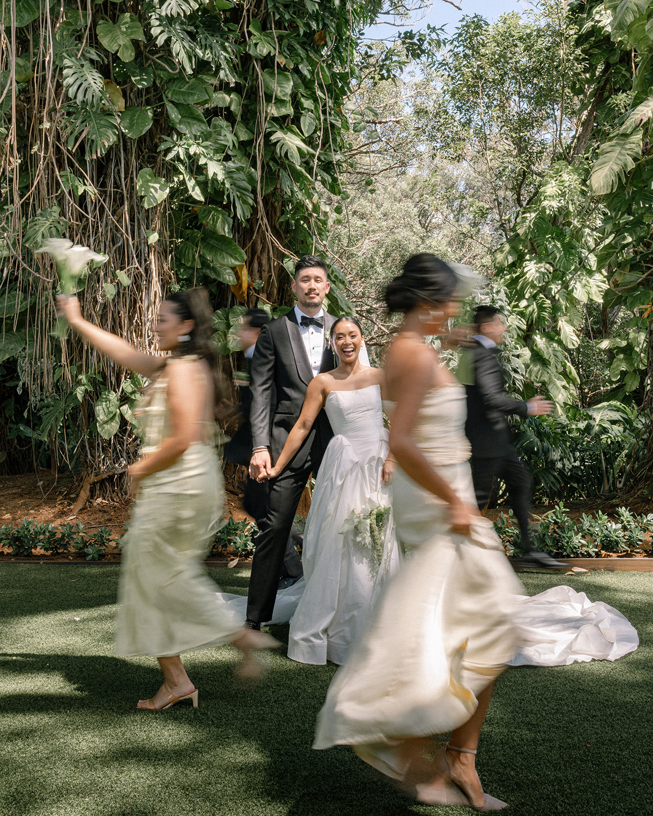 A joyful couple with their wedding party at Sunset Ranch Oahu, surrounded by lush greenery. The bride smiles brightly as her bridesmaids walk past, creating a lively and vibrant moment.