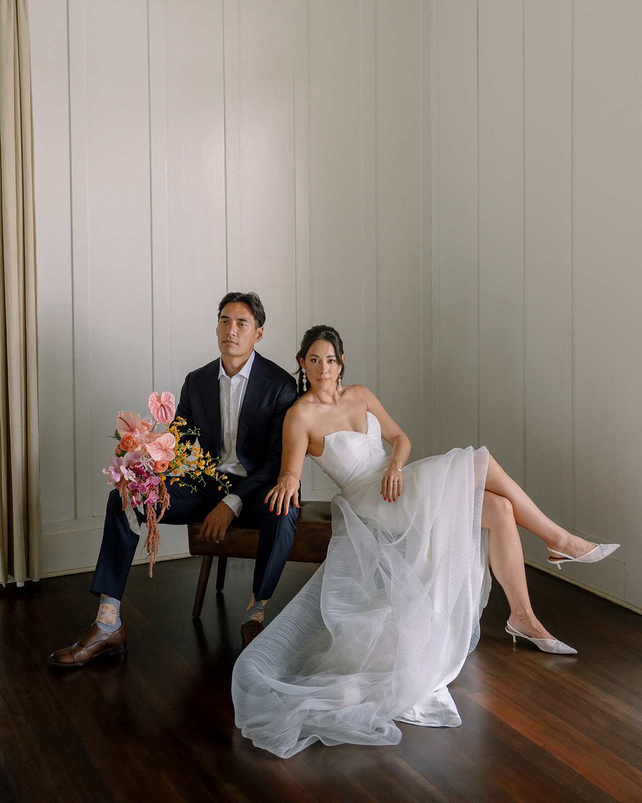 Couple seated elegantly for their Dillingham Ranch Wedding, with the bride holding a vibrant bouquet and wearing a flowing, textured wedding gown while the groom stands beside her in a tailored suit, set against a minimalist background.