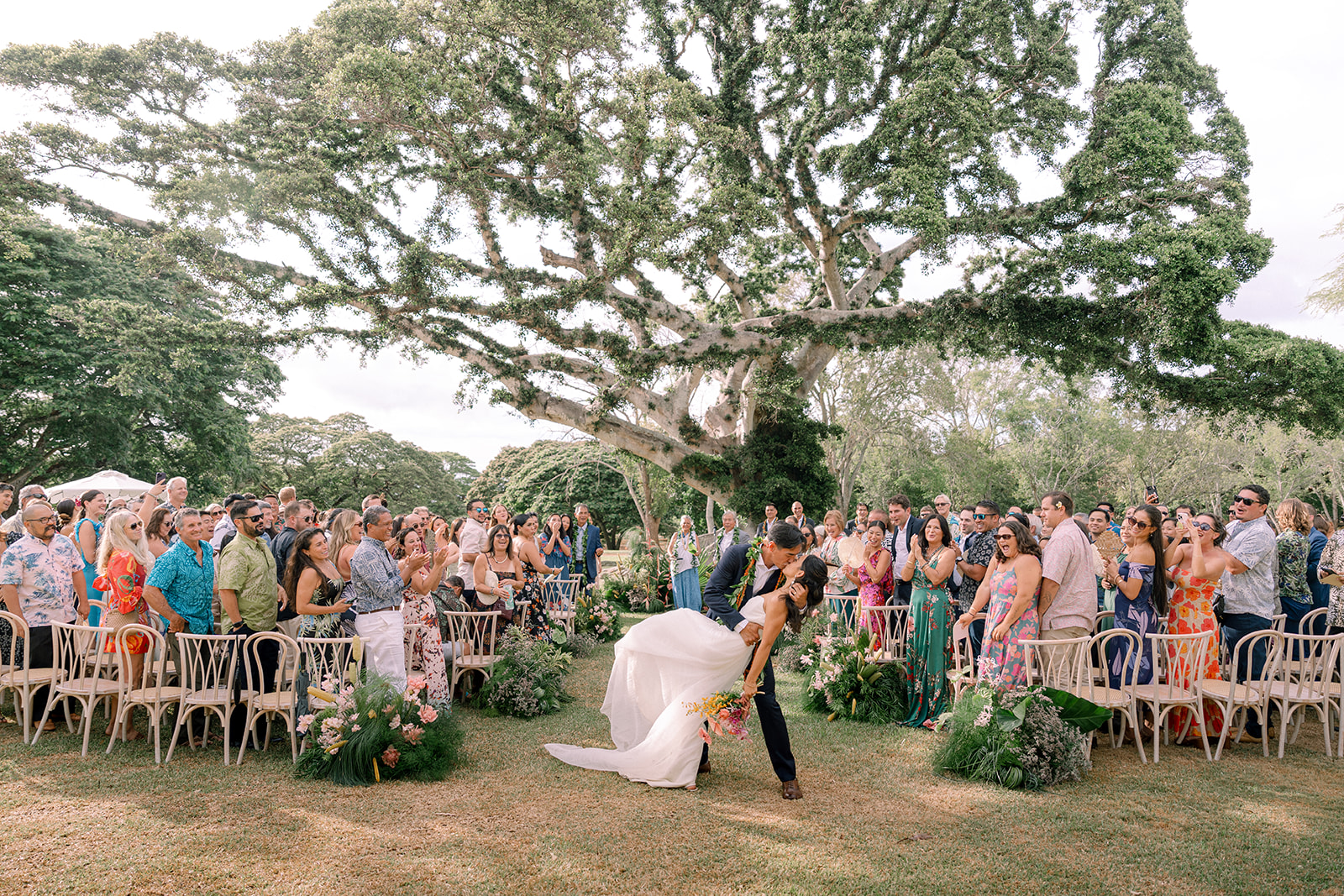Bride and groom kiss under a banyan tree at their Dillingham Ranch wedding, surrounded by cheering guests on the North Shore of Oahu.
