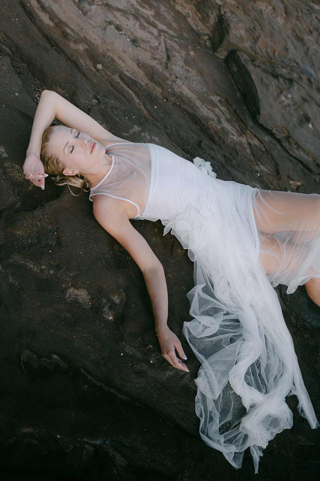 A bride in a flowing white bridal dress lies on the rocks at an Oahu beach, showcasing effortless editorial imagery with natural beauty and elegance.