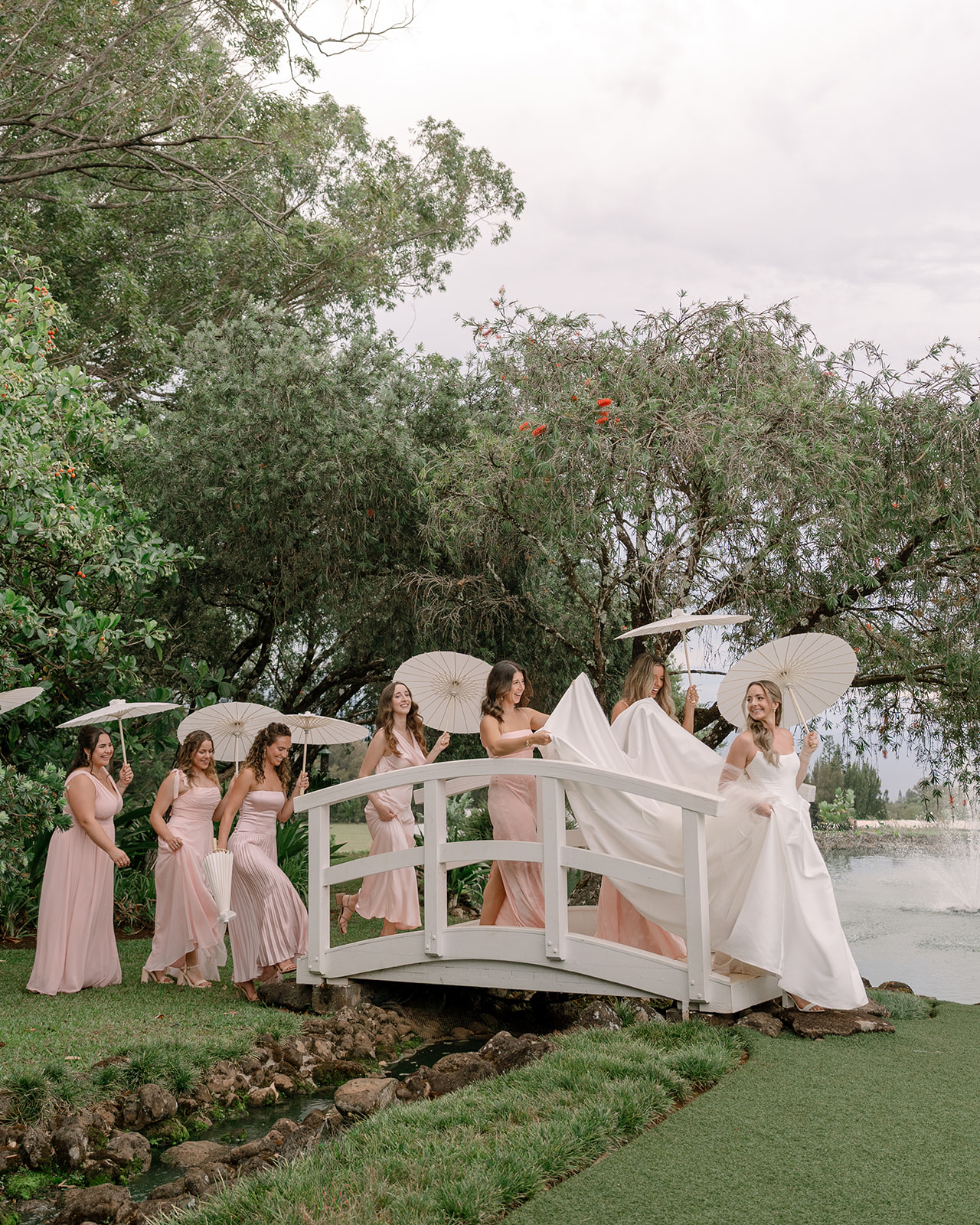 Bridesmaids walking over a bridge with parasols at a multi-day wedding in Hawaii, captured during a beautiful outdoor ceremony at Sunset Ranch Oahu.