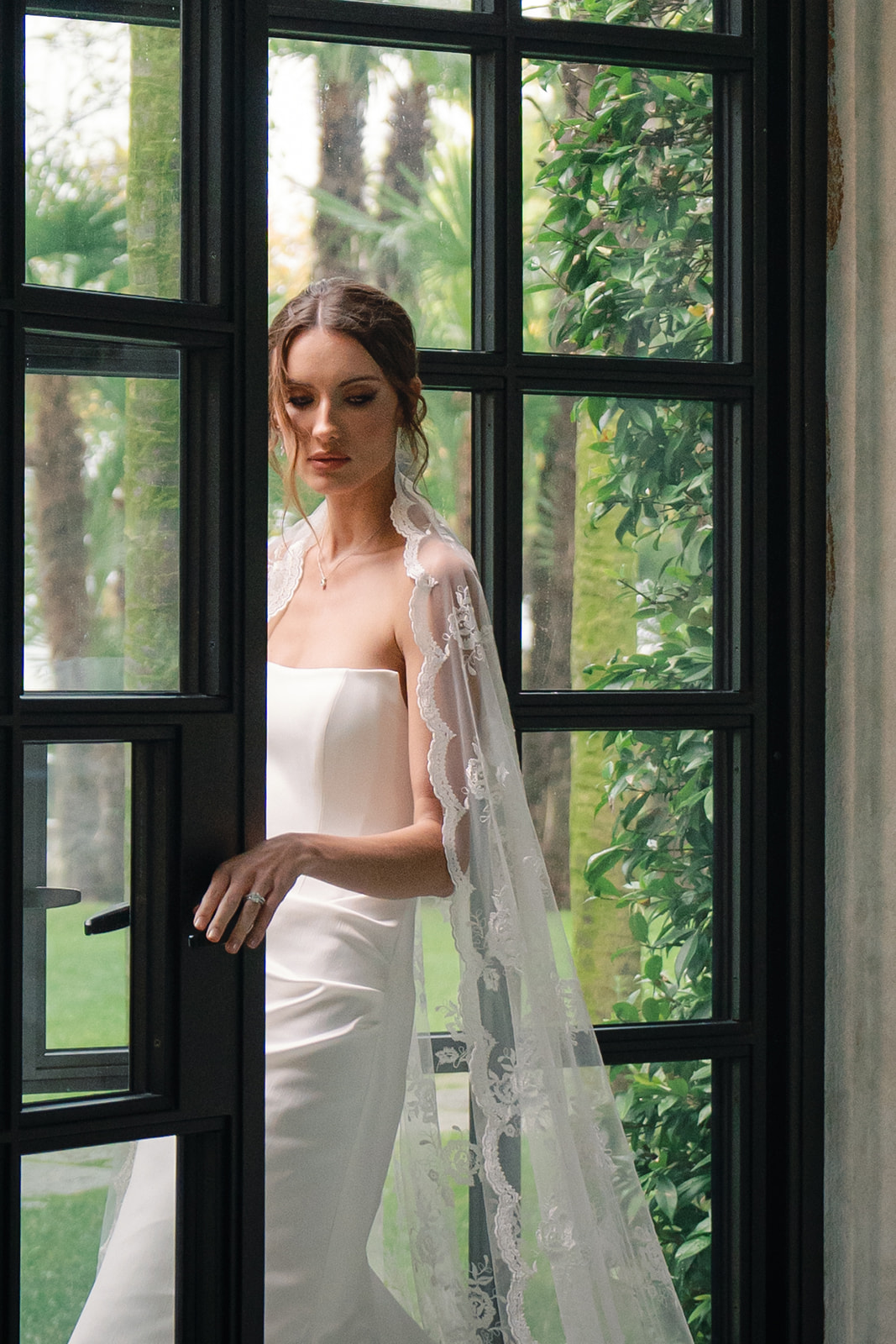 Bride in a lace veil holding the door handle, standing gracefully by a window at a villa with greenery outside. Evolution of wedding photography captured in this timeless moment.