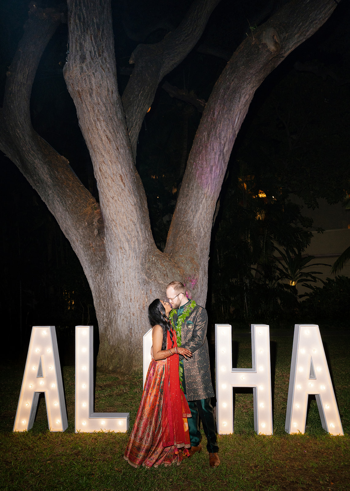 Couple in vibrant Indian attire kissing by lit “ALOHA” marquee under a tree during their Sangeet at the Royal Hawaiian Hotel Waikiki.