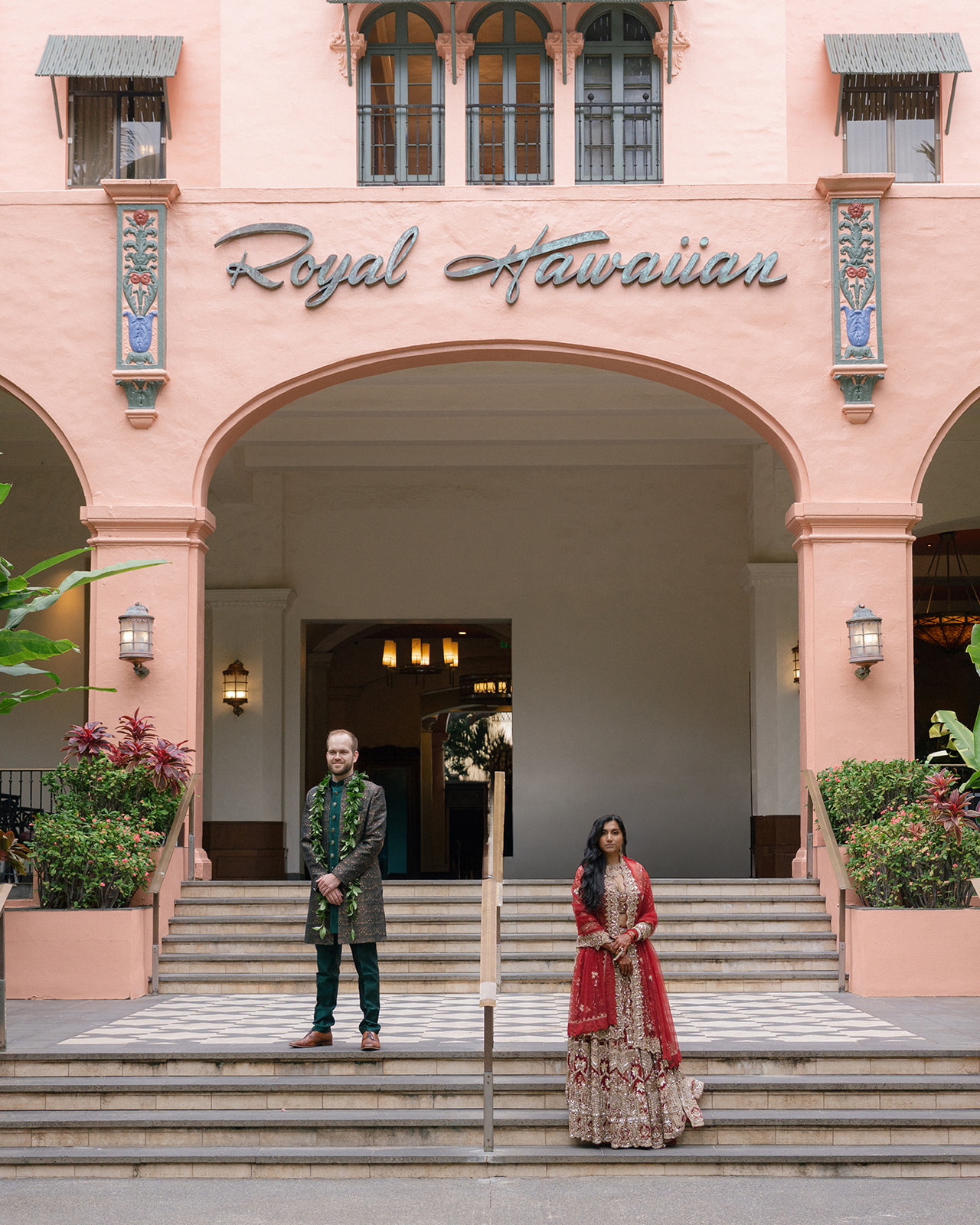 Couple in traditional Indian wedding attire standing on the front steps of Royal Hawaiian Hotel Waikiki beneath the iconic pink archway.