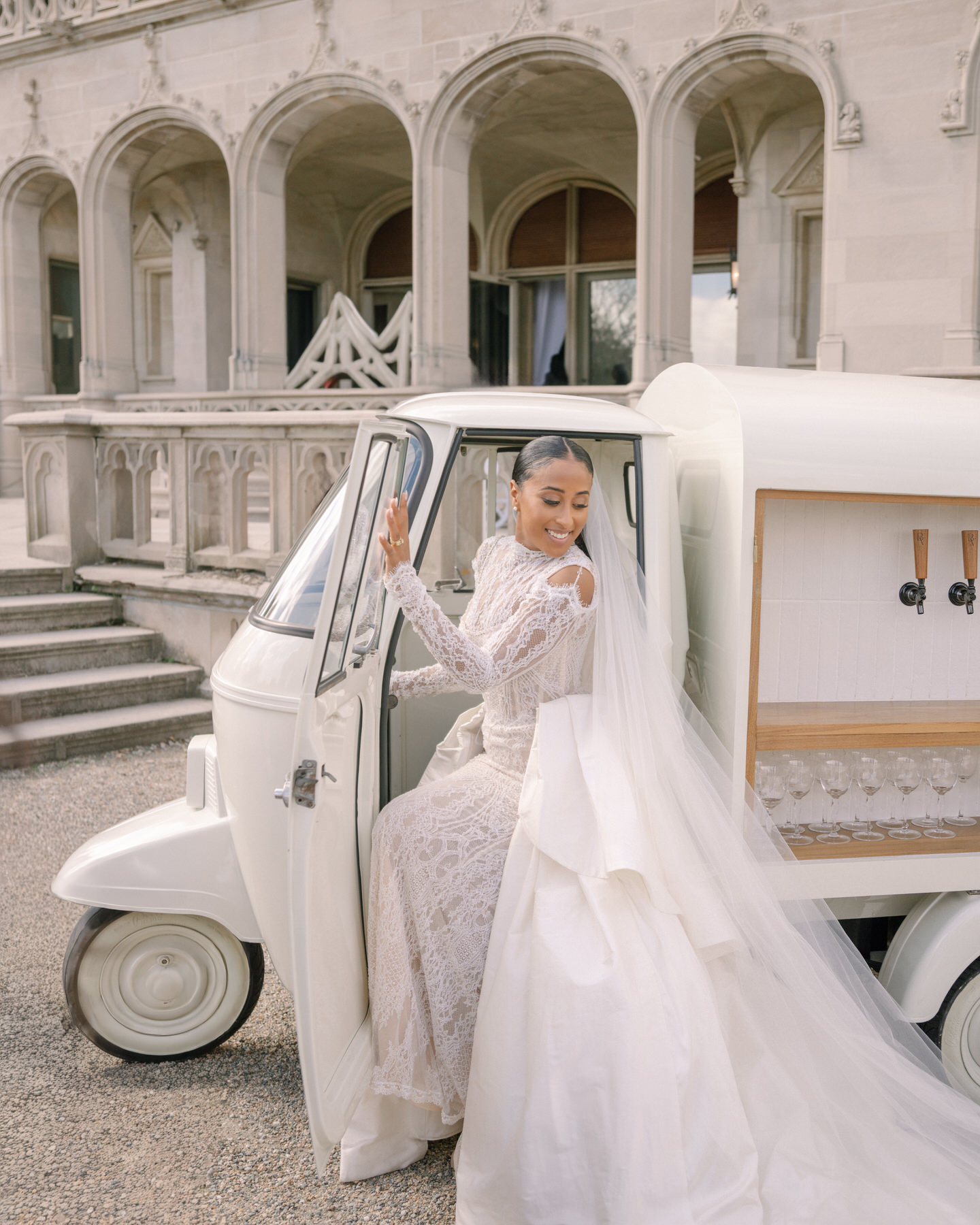 Bride smiling as she exits a classic vehicle at the brunch wedding at Ochre Court Newport, her elegant lace wedding gown flowing behind her.
