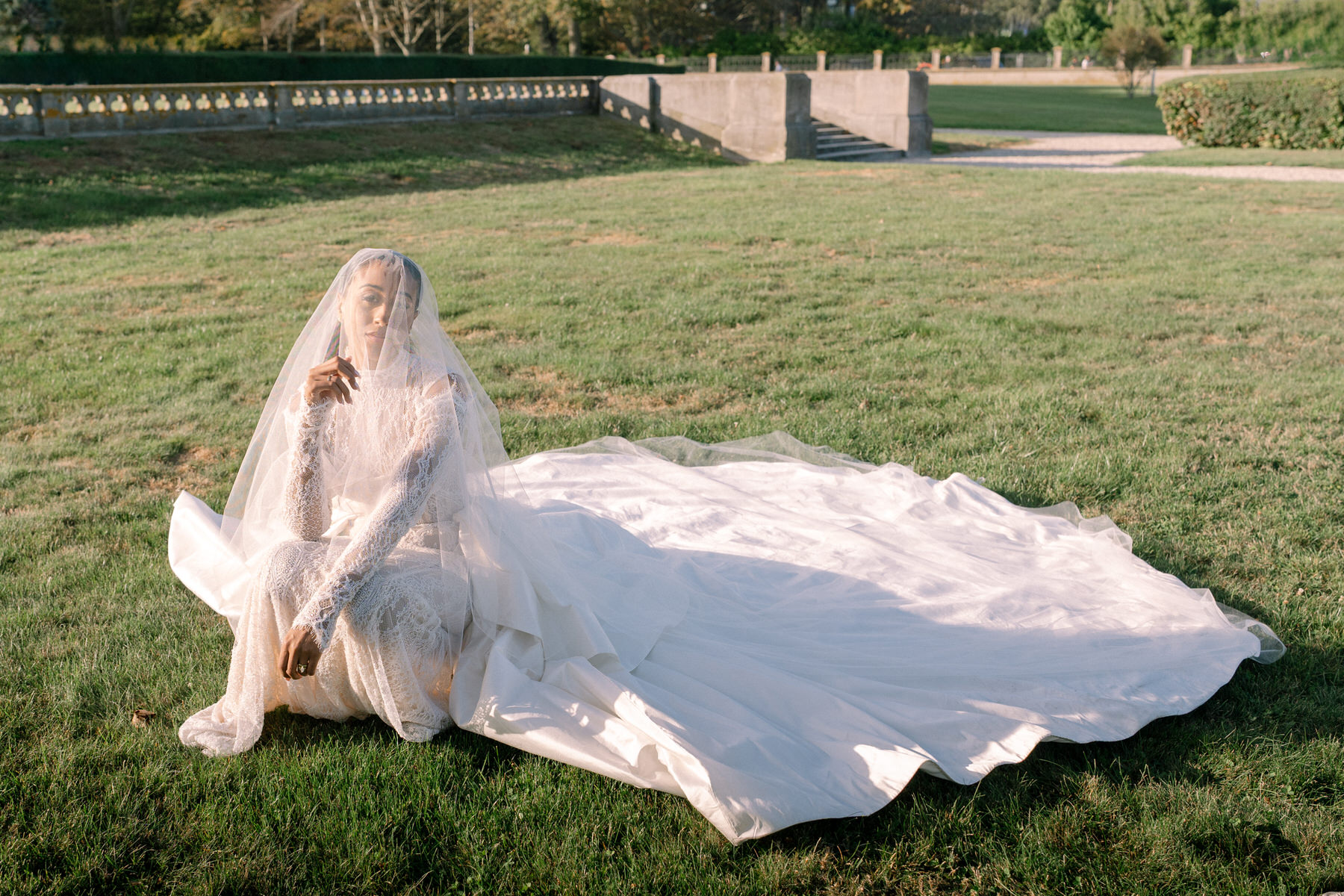 Bride seated on the grass with her flowing wedding gown and veil trailing behind her, showcasing the elegance of her wedding attire.
