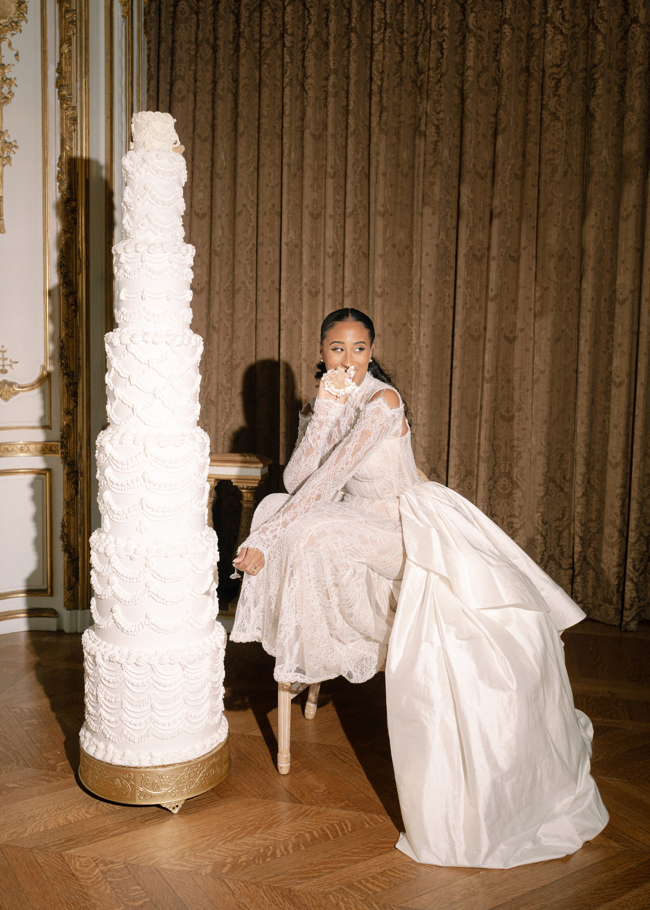 Bride sitting gracefully beside the towering wedding cake, showcasing her elegant wedding dress in a classic moment.
