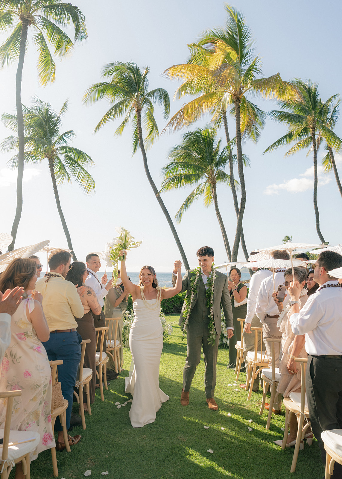Newlyweds raise their joined hands while walking up the aisle after their outdoor Lanikuhonua wedding ceremony