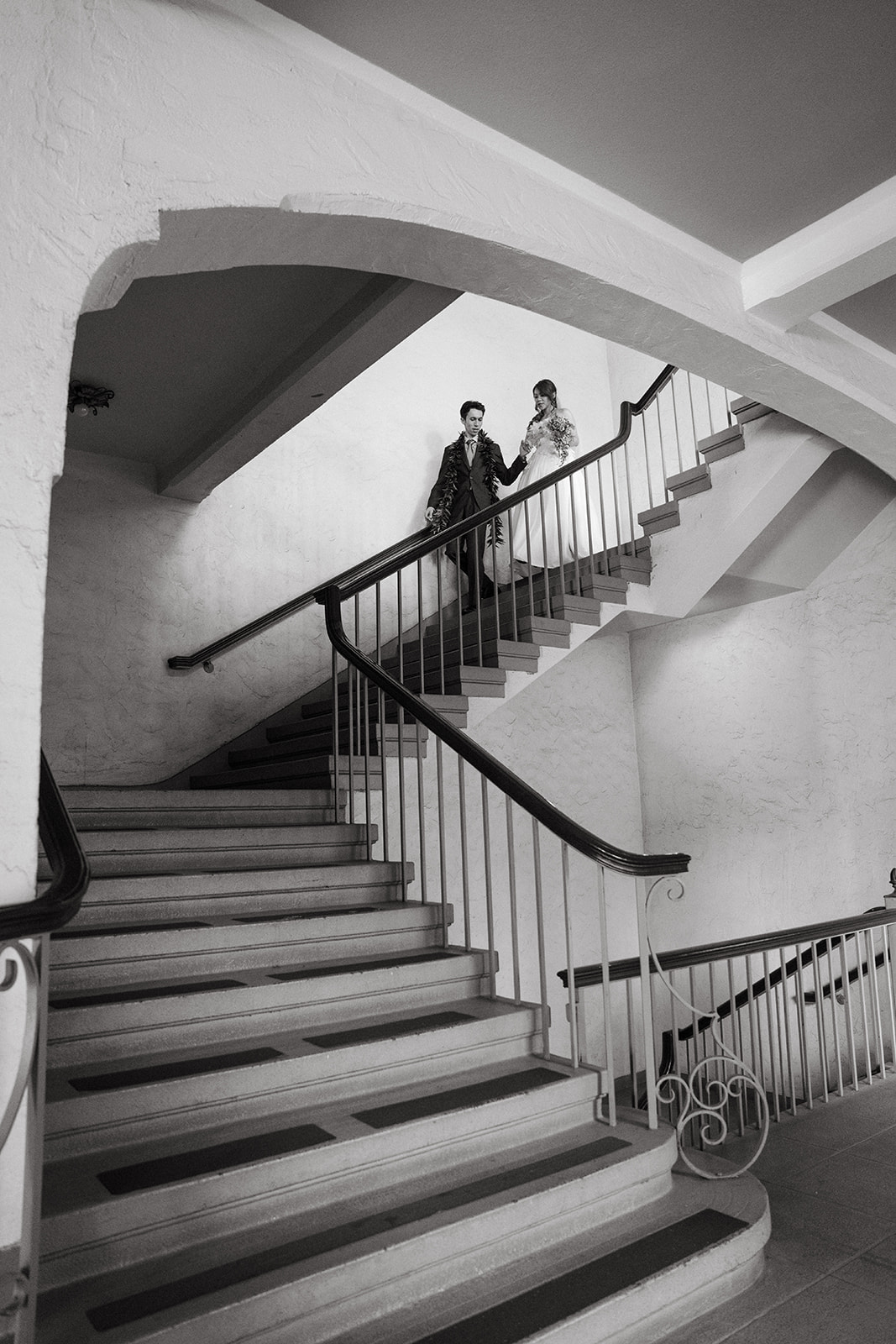Black-and-white wedding portrait of a couple standing on the grand staircase inside ʻIolani Palace.
