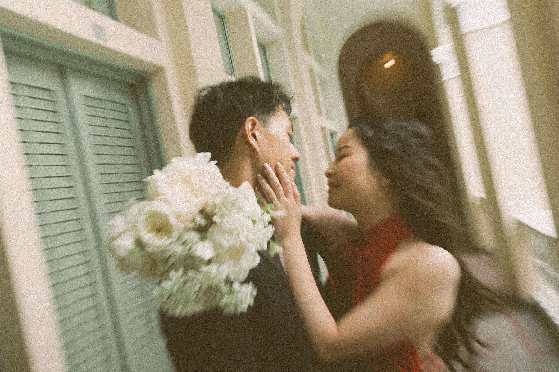 Bride in a red dress touches the groom’s face as he holds a white bouquet in an arched corridor at Cafe Julia.