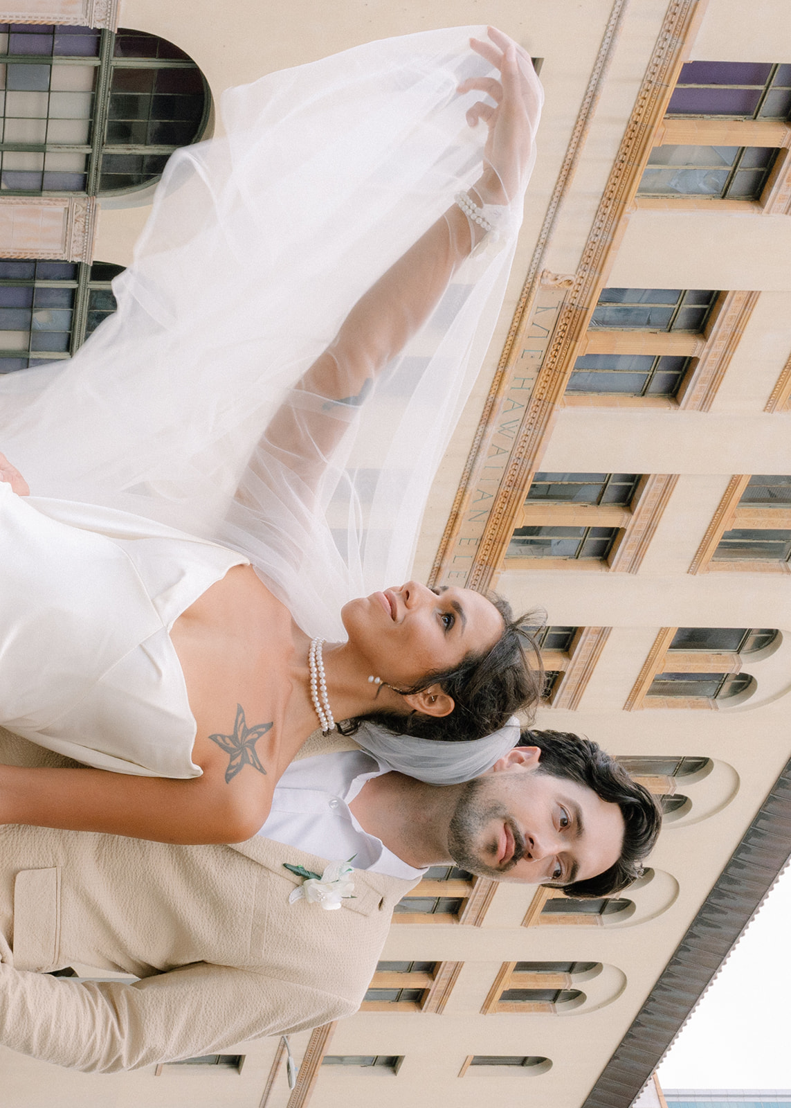 Bride lifts her flowing veil beside the groom in a beige suit during a modern Honolulu wedding portrait outside the Hawaii Theatre.