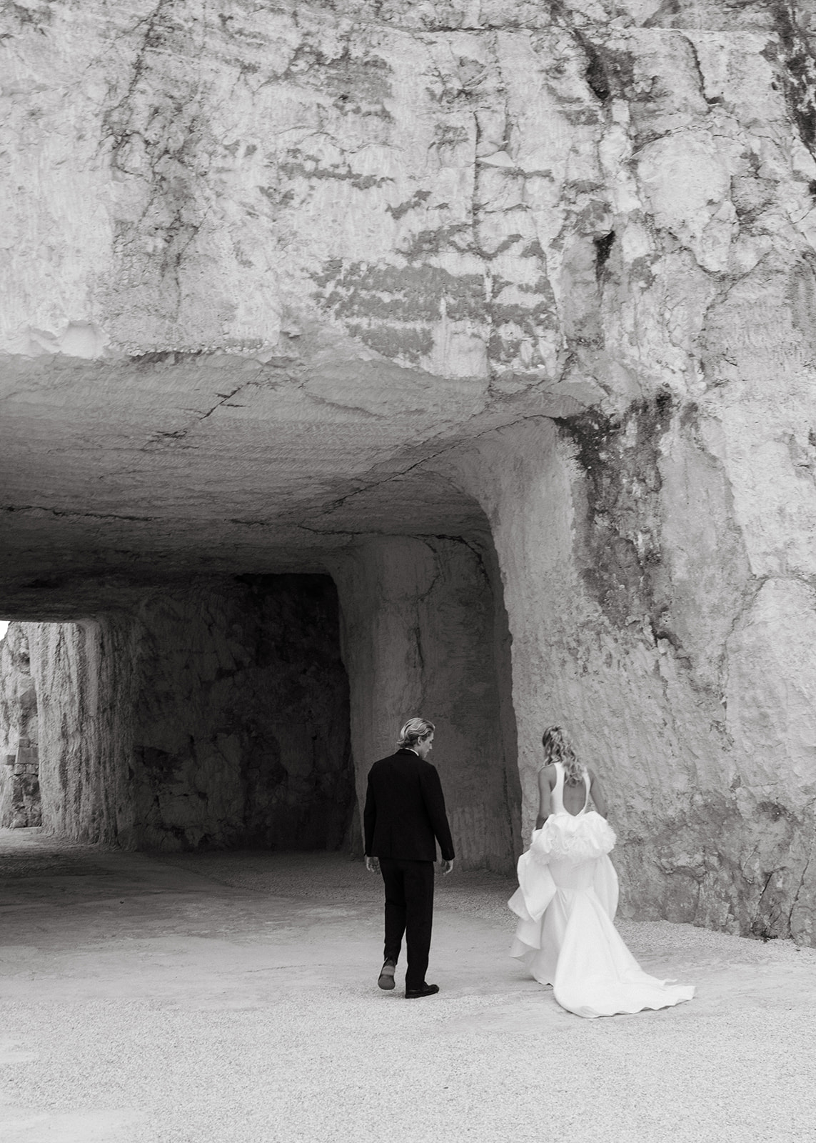 Bride and groom walk beneath a monumental carved-stone passage at Cava Puglia.