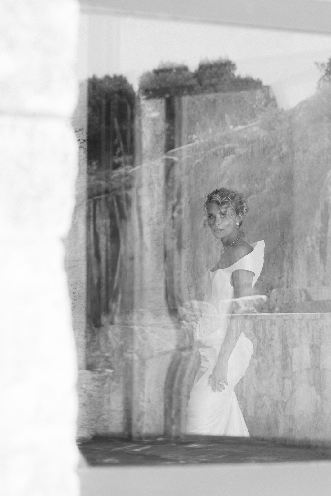 Black-and-white portrait of a bride in a white gown seen through reflective glass at Cava Puglia.