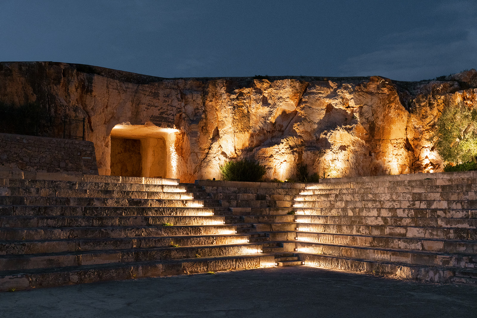 Illuminated limestone walls and tiered amphitheatre seating at Cava Puglia after dark.
