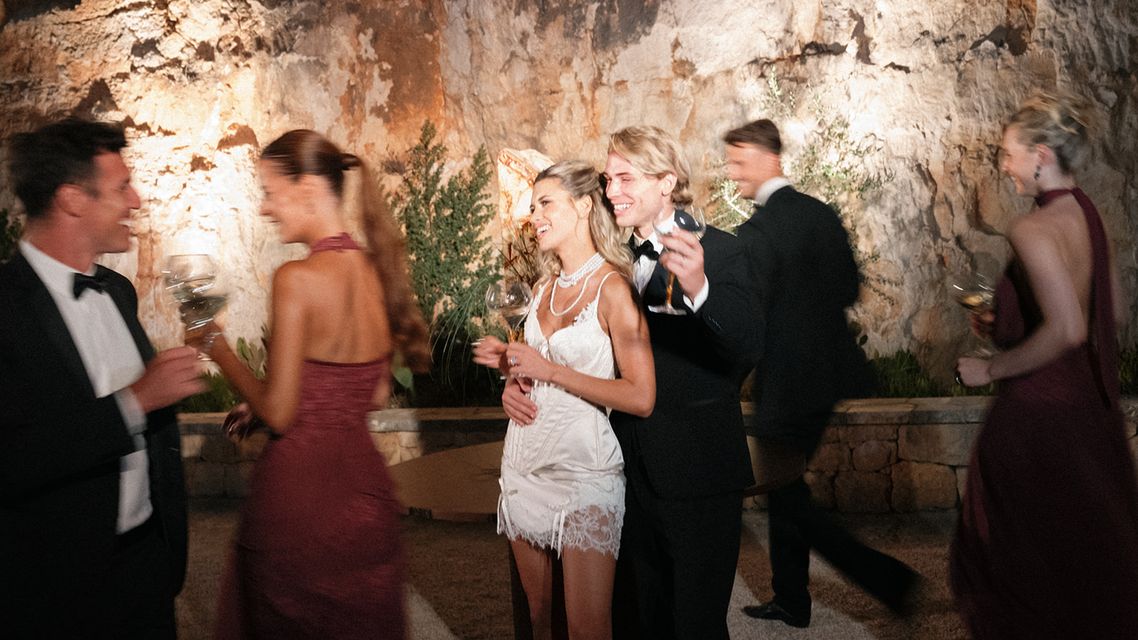 Bride and groom hold wine glasses as guests mingle beside illuminated limestone walls at Cava Puglia.