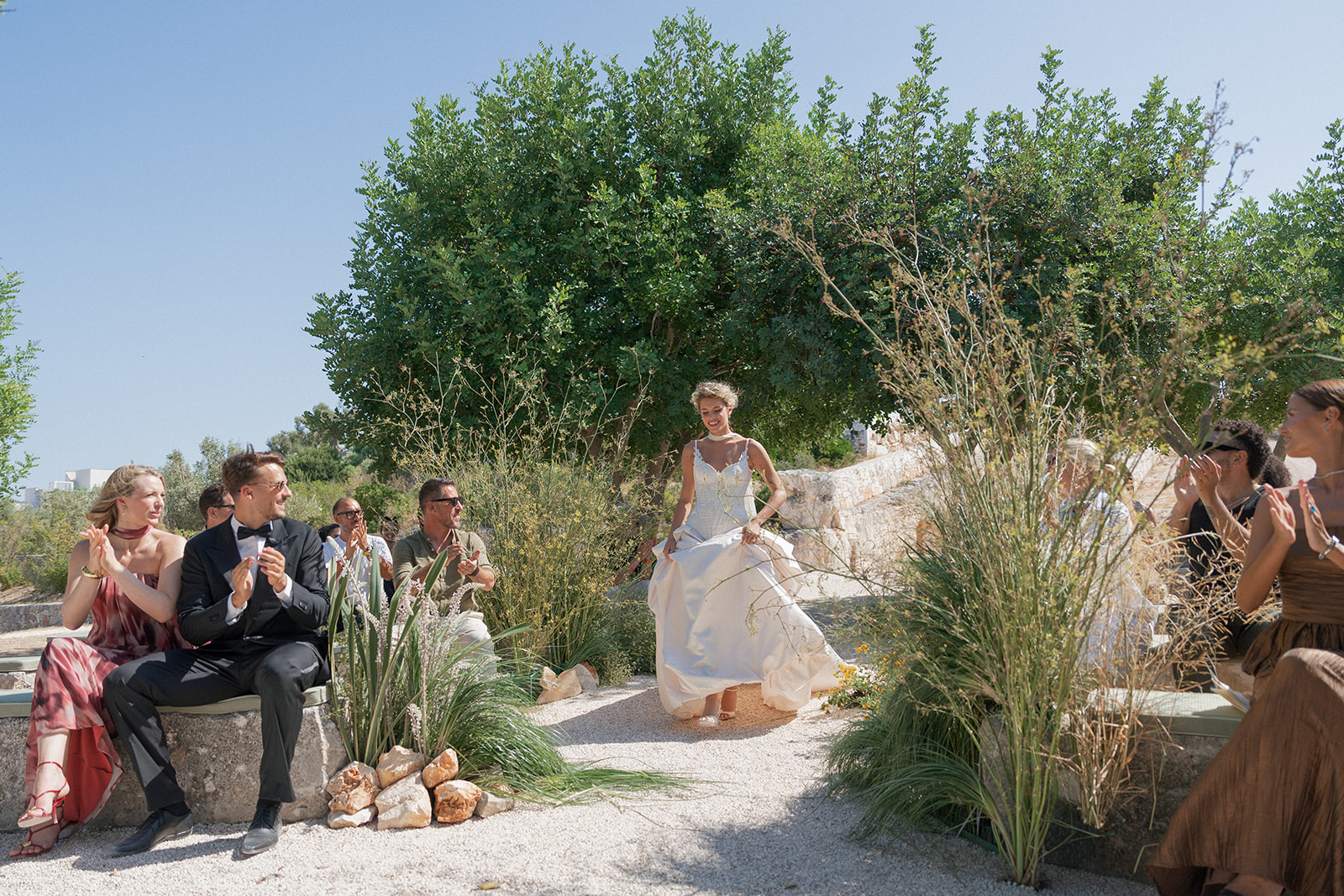 Bride walks down a gravel ceremony aisle framed by grasses and citrus trees at Cava Puglia.