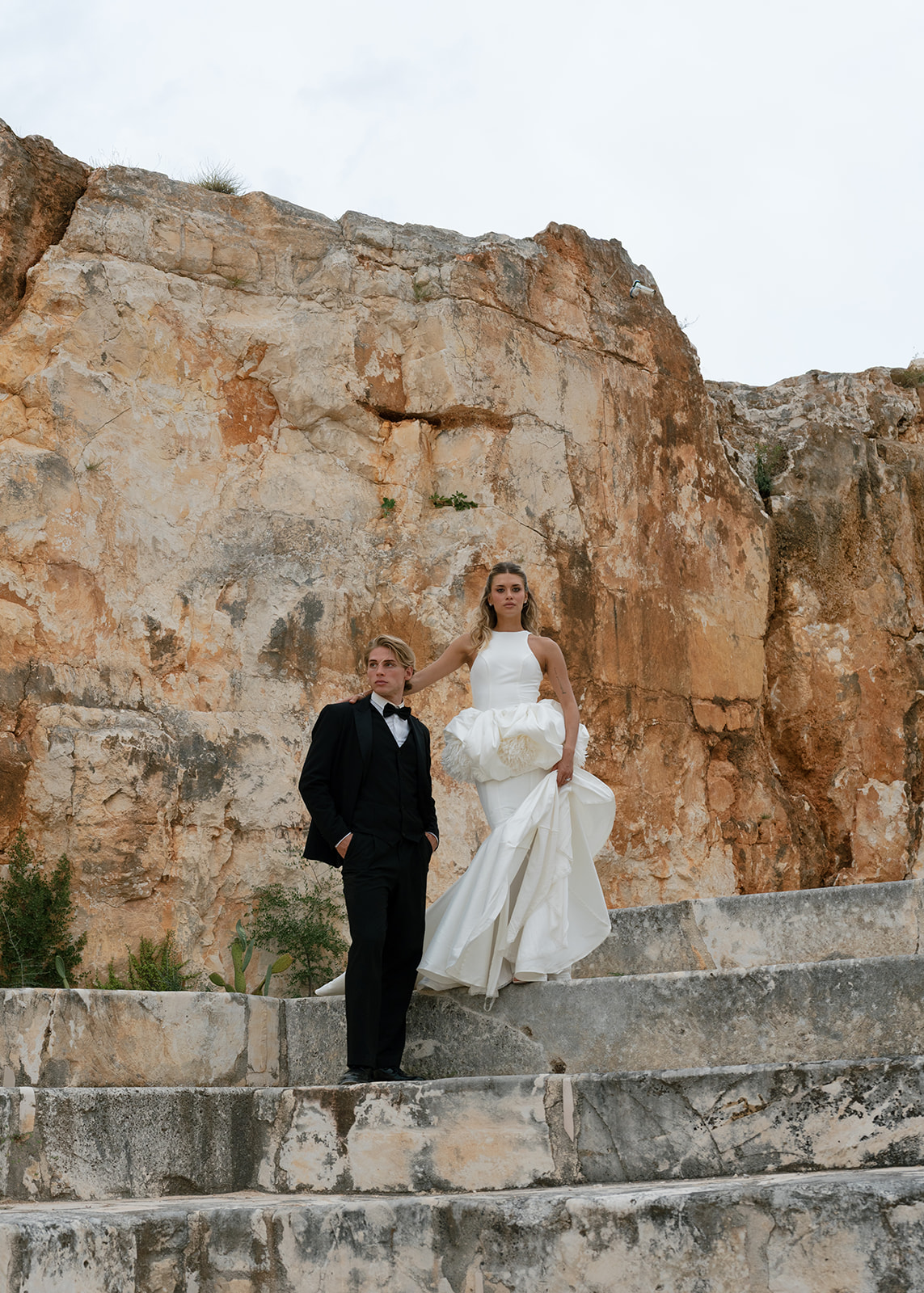 Bride in a sculptural white gown and groom in a black tuxedo pose against Cava Puglia’s limestone walls for an Italy wedding.