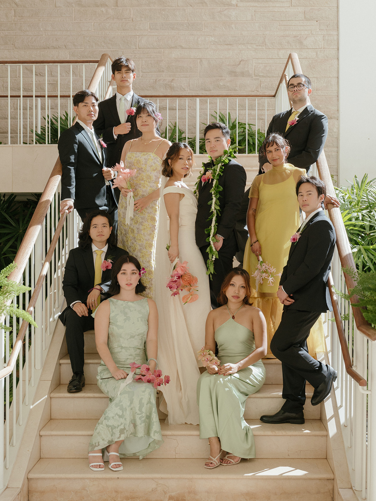 Couple and wedding party on a staircase, with black suits, sage and yellow dresses, and pink orchid bouquets.