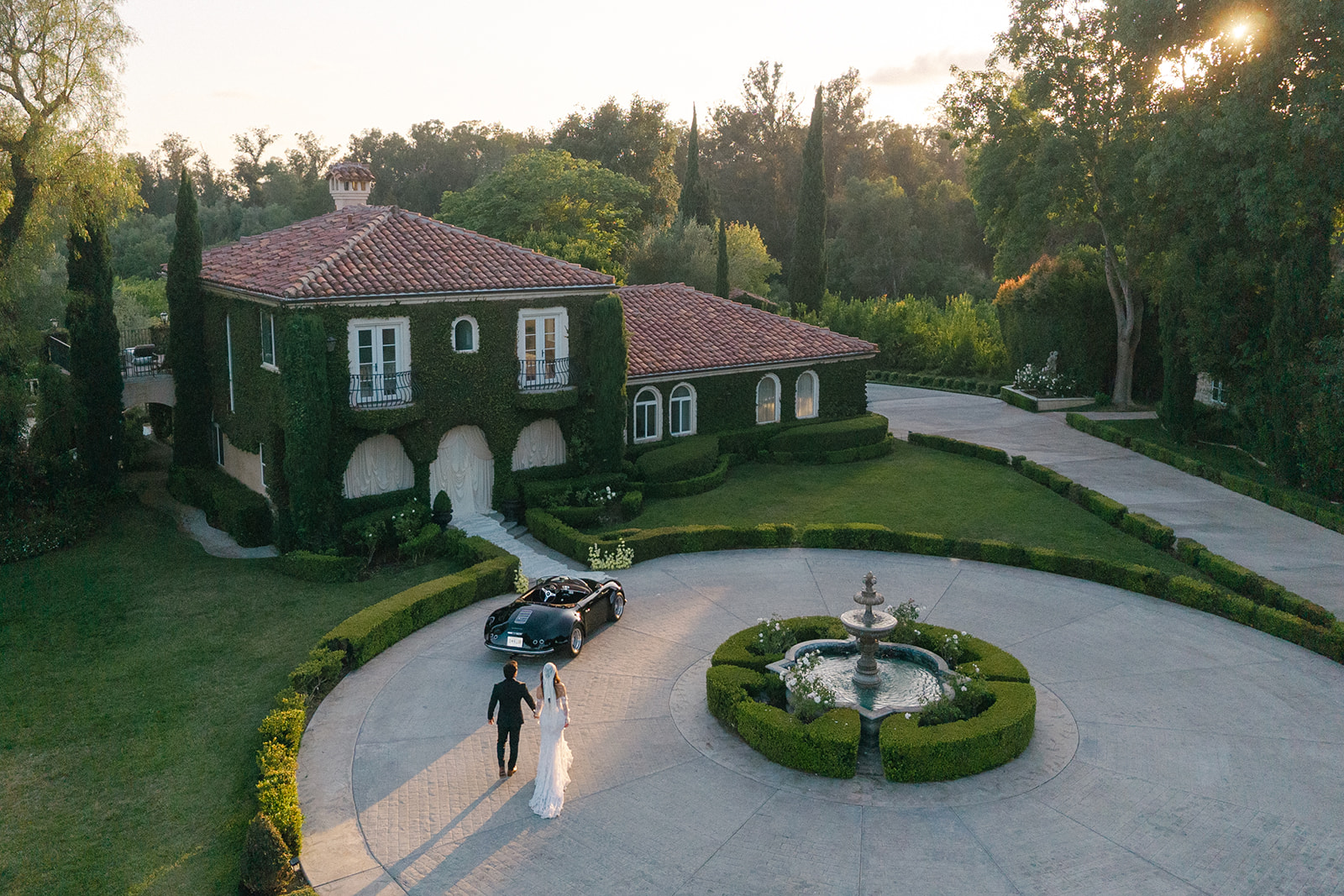 Aerial view of Villa Amodei with a circular fountain, ivy-covered walls, and a couple walking toward a convertible.