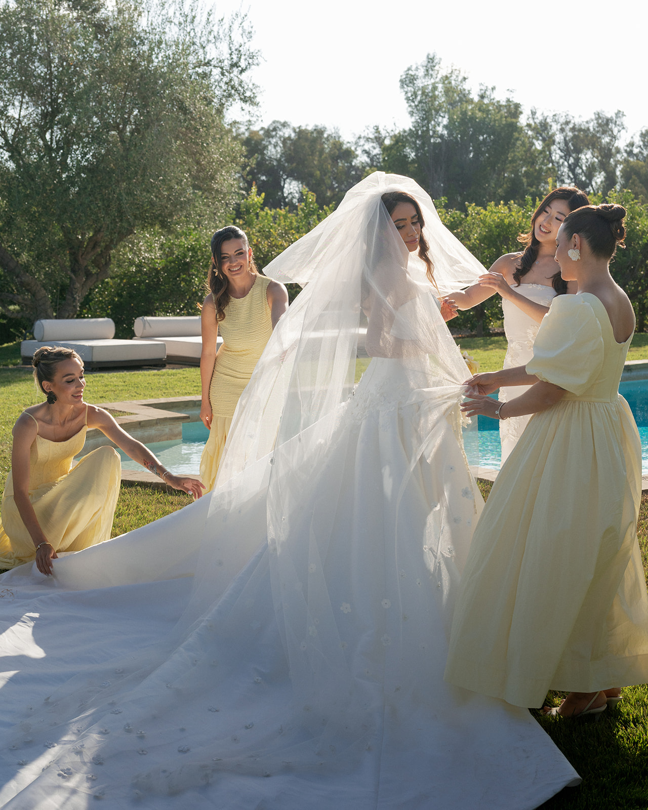 Bridesmaids in yellow dresses arrange the bride’s veil beside the pool at Villa Amodei.