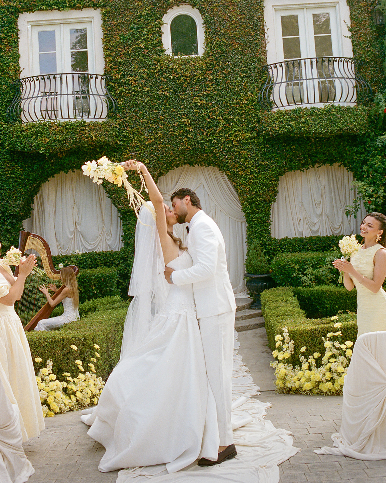 Bride raises her bouquet while kissing the groom outside Villa Amodei as bridesmaids applaud.