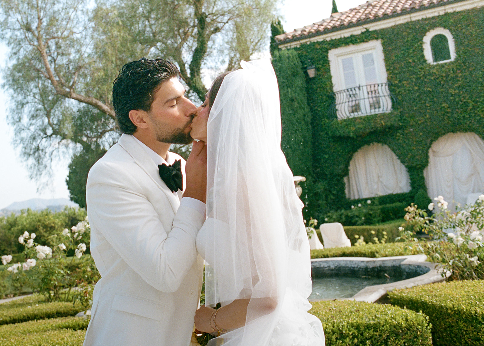 Bride and groom kiss beside the garden fountain with Villa Amodei’s ivy-covered facade behind them.